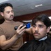 A barber is cutting the hair of a seated client in what appears to be a barbershop. The barber, wearing a brown t-shirt, is using electric clippers with a blue guard attachment on the client's hair. The client is draped in a black barber cape and appears to be looking forward calmly.