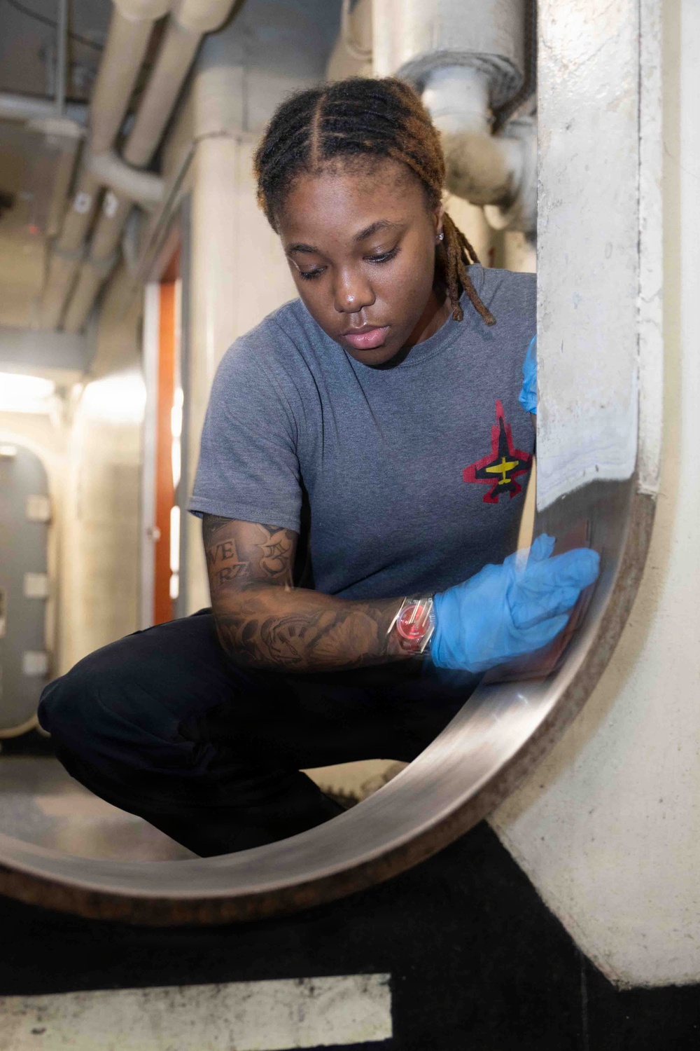 A young woman wearing blue nitrile gloves and a gray t-shirt with a military aircraft insignia is crouched down performing maintenance or cleaning work on a large cylindrical metal component. The setting appears to be an industrial or military vessel interior, evidenced by the overhead piping, confined space, and institutional infrastructure visible in the background. She is focused intently on her task, working near the base or rim of a large stainless steel vessel or hatch.