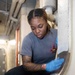A young woman wearing blue nitrile gloves and a gray t-shirt with a military aircraft insignia is crouched down performing maintenance or cleaning work on a large cylindrical metal component. The setting appears to be an industrial or military vessel interior, evidenced by the overhead piping, confined space, and institutional infrastructure visible in the background. She is focused intently on her task, working near the base or rim of a large stainless steel vessel or hatch.