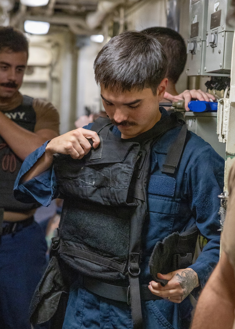A young male in a blue naval coverall uniform is donning or adjusting a black tactical body armor/plate carrier vest in a narrow shipboard corridor. Several other personnel are visible in the background, one wearing a shirt with 'NAVY' text. The setting appears to be the interior passageway of a military vessel.