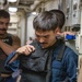 A young male in a blue naval coverall uniform is donning or adjusting a black tactical body armor/plate carrier vest in a narrow shipboard corridor. Several other personnel are visible in the background, one wearing a shirt with 'NAVY' text. The setting appears to be the interior passageway of a military vessel.