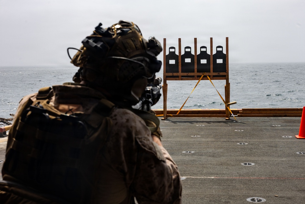 A heavily equipped military personnel is conducting a live-fire marksmanship training exercise on what appears to be the deck of a naval vessel at sea. The shooter is aiming at a wooden target stand holding four standard silhouette shooting targets. The overcast sky and open ocean are visible in the background, confirming an at-sea training environment.