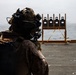 A heavily equipped military personnel is conducting a live-fire marksmanship training exercise on what appears to be the deck of a naval vessel at sea. The shooter is aiming at a wooden target stand holding four standard silhouette shooting targets. The overcast sky and open ocean are visible in the background, confirming an at-sea training environment.