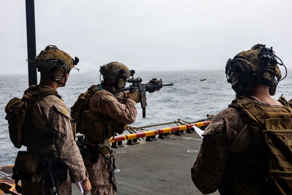 Three military personnel in full combat gear are positioned on the deck of a naval vessel, with one actively firing a rifle toward the open ocean. The soldiers are wearing desert camouflage uniforms, tactical vests, and helmets equipped with night-vision device mounts. This appears to be a live-fire training exercise conducted from a ship at sea.