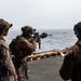 Three military personnel in full combat gear are positioned on the deck of a naval vessel, with one actively firing a rifle toward the open ocean. The soldiers are wearing desert camouflage uniforms, tactical vests, and helmets equipped with night-vision device mounts. This appears to be a live-fire training exercise conducted from a ship at sea.