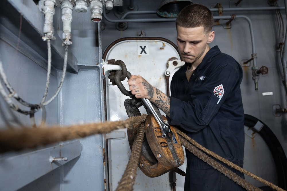A U.S. Navy sailor in dark blue coveralls is working with rigging hardware aboard a naval vessel. He is using a wrench or tool to work on a shackle connected to a block-and-tackle pulley system with heavy rope. The setting appears to be an interior or sheltered deck area of a ship, identifiable by the gray painted steel bulkheads and piping.
