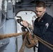 A U.S. Navy sailor in dark blue coveralls is working with rigging hardware aboard a naval vessel. He is using a wrench or tool to work on a shackle connected to a block-and-tackle pulley system with heavy rope. The setting appears to be an interior or sheltered deck area of a ship, identifiable by the gray painted steel bulkheads and piping.