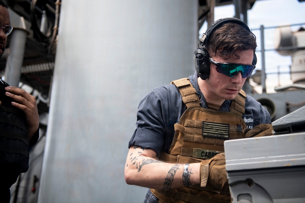 A military or naval personnel member wearing a tan plate carrier vest, tactical headset, and tinted safety glasses is focused on operating equipment aboard what appears to be a naval vessel. A name tape reading 'CABRER' (partially visible) is affixed to the vest. A second individual is partially visible on the left side of the frame.