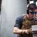 A military or naval personnel member wearing a tan plate carrier vest, tactical headset, and tinted safety glasses is focused on operating equipment aboard what appears to be a naval vessel. A name tape reading 'CABRER' (partially visible) is affixed to the vest. A second individual is partially visible on the left side of the frame.
