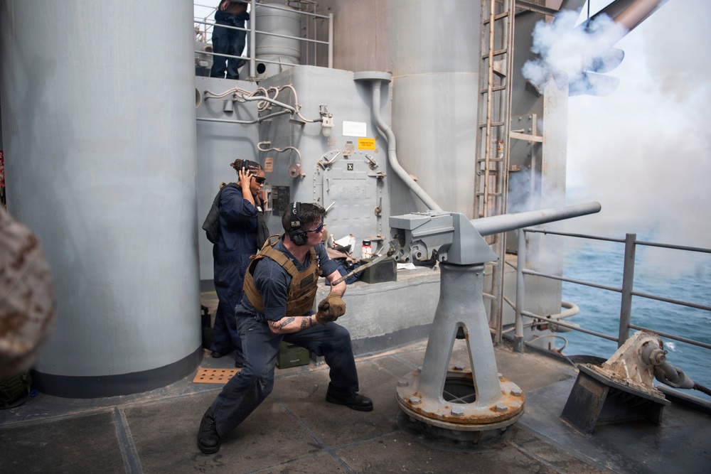 Two naval personnel are operating a deck-mounted gun on a warship, with visible smoke and a muzzle blast indicating the weapon has just been fired. One sailor in a tactical vest and hearing protection is crouched at the gun mount, while a second sailor stands nearby covering their ears. The scene takes place on the exterior deck of a gray military vessel with ocean visible in the background.