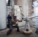 Two naval personnel are operating a deck-mounted gun on a warship, with visible smoke and a muzzle blast indicating the weapon has just been fired. One sailor in a tactical vest and hearing protection is crouched at the gun mount, while a second sailor stands nearby covering their ears. The scene takes place on the exterior deck of a gray military vessel with ocean visible in the background.