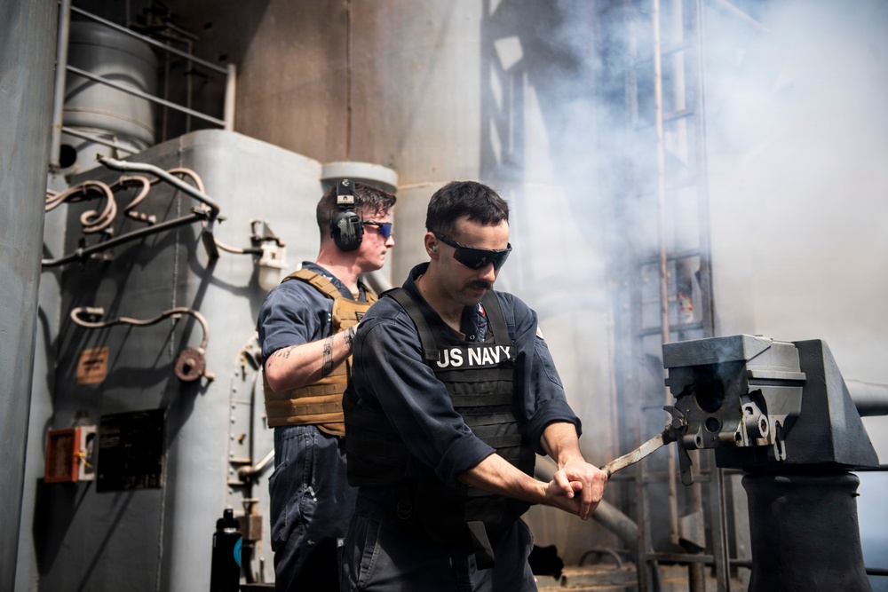 Two U.S. Navy sailors are operating on the deck of a naval vessel amid visible smoke or gun discharge residue. The foreground sailor wears a 'US NAVY' labeled vest and sunglasses while handling what appears to be a weapon system component. The background sailor wears hearing protection (earmuffs) and a tan tactical vest.
