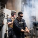 Two U.S. Navy sailors are operating on the deck of a naval vessel amid visible smoke or gun discharge residue. The foreground sailor wears a 'US NAVY' labeled vest and sunglasses while handling what appears to be a weapon system component. The background sailor wears hearing protection (earmuffs) and a tan tactical vest.