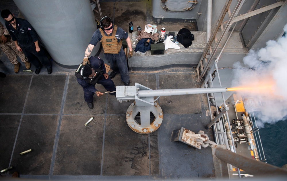 An overhead view of a naval vessel deck showing military personnel conducting a live-fire exercise with a deck-mounted cannon. The weapon has just been fired, as evidenced by the visible muzzle flash and smoke emanating from the barrel. Spent shell casings are visible on the deck, and personnel are wearing hearing protection and tactical gear.