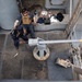 An overhead view of a naval vessel deck showing military personnel conducting a live-fire exercise with a deck-mounted cannon. The weapon has just been fired, as evidenced by the visible muzzle flash and smoke emanating from the barrel. Spent shell casings are visible on the deck, and personnel are wearing hearing protection and tactical gear.