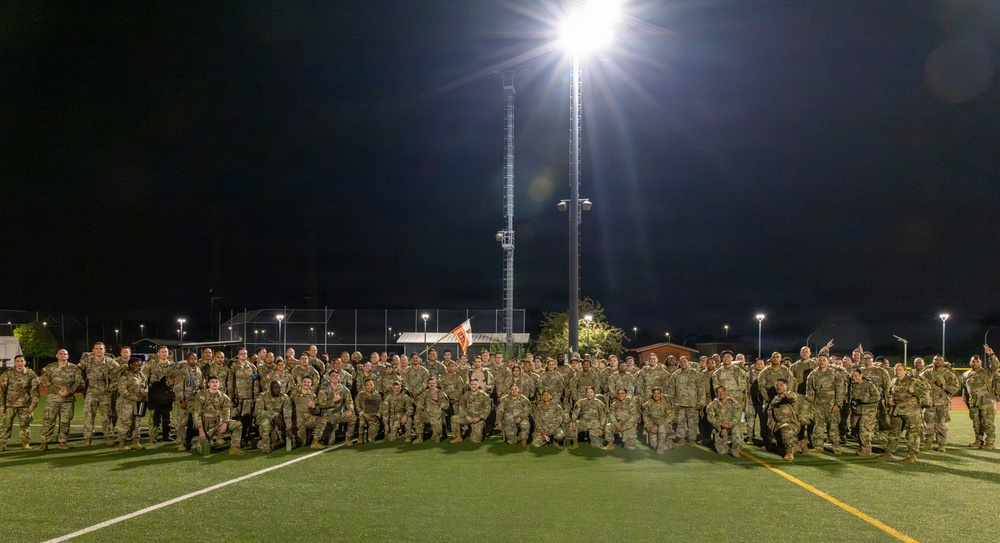 A large group of military personnel in camouflage uniforms are posed for a group photograph on an illuminated athletic field at night. The soldiers are arranged in multiple rows, with some kneeling in front and others standing behind, suggesting a formal unit photo. The scene is lit by tall stadium-style light poles, with what appears to be a military installation or sports complex in the background.