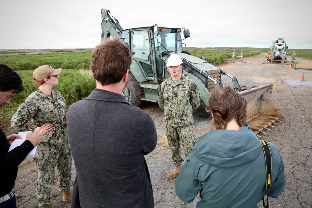 A U.S. Navy service member wearing a white hard hat is being interviewed or briefed by civilians, including a photographer with a Nikon camera, at an active construction or runway repair site. Another Navy service member in camouflage uniform with a tan cap is also present, taking notes. In the background, additional personnel and heavy construction equipment, including a concrete mixer truck, are visible on what appears to be a paved surface under repair.