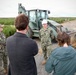 A U.S. Navy service member wearing a white hard hat is being interviewed or briefed by civilians, including a photographer with a Nikon camera, at an active construction or runway repair site. Another Navy service member in camouflage uniform with a tan cap is also present, taking notes. In the background, additional personnel and heavy construction equipment, including a concrete mixer truck, are visible on what appears to be a paved surface under repair.
