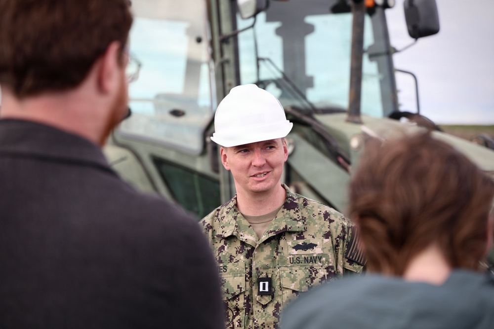 A U.S. Navy officer wearing NWU (Navy Working Uniform) camouflage and a white hard hat is speaking to a small group of civilians or personnel in the foreground. The officer displays rank insignia consistent with a Lieutenant (O-3) and a warfare qualification badge on the uniform. The setting appears to be an outdoor construction or equipment site, with large military or heavy machinery visible in the background.