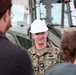 A U.S. Navy officer wearing NWU (Navy Working Uniform) camouflage and a white hard hat is speaking to a small group of civilians or personnel in the foreground. The officer displays rank insignia consistent with a Lieutenant (O-3) and a warfare qualification badge on the uniform. The setting appears to be an outdoor construction or equipment site, with large military or heavy machinery visible in the background.