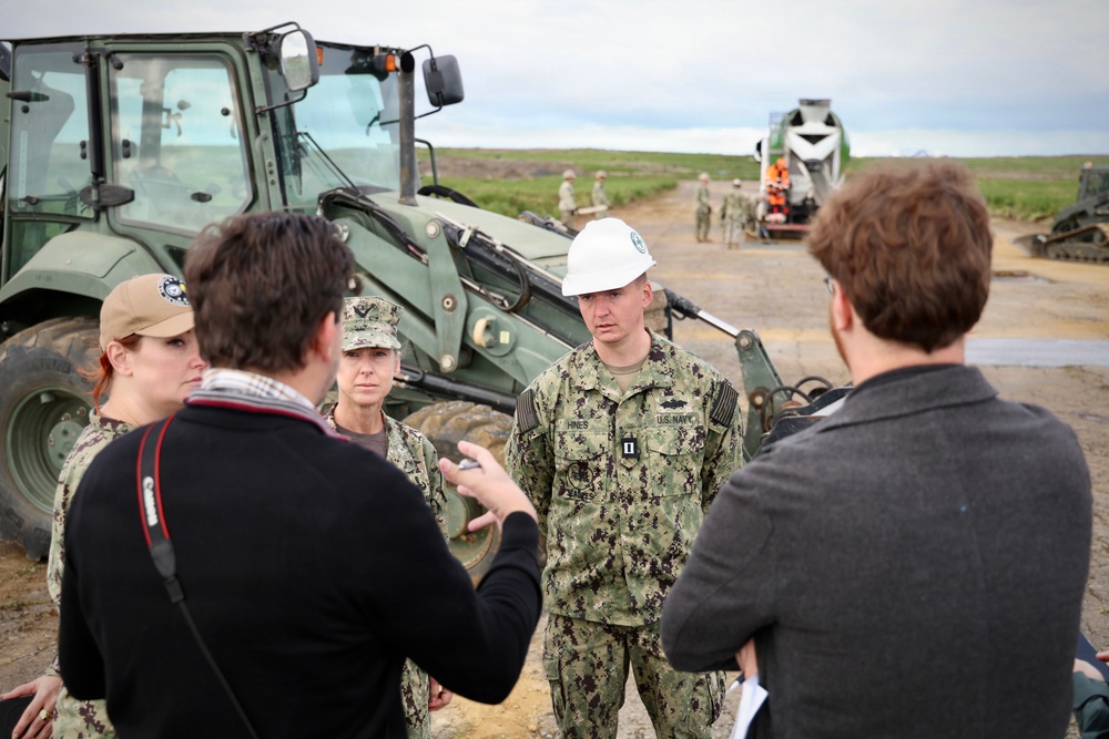 A group of U.S. Navy personnel in camouflage uniforms are engaged in a briefing or discussion with two civilians at an outdoor construction or engineering site. One Navy officer wearing a white hard hat has a name tape reading 'HINES' and rank insignia indicating a Captain (O-6) or Lieutenant, with 'U.S. NAVY' clearly visible on the uniform. Military construction equipment including a front-end loader and what appears to be a concrete mixer are visible in the background along with additional personnel.