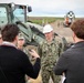 A group of U.S. Navy personnel in camouflage uniforms are engaged in a briefing or discussion with two civilians at an outdoor construction or engineering site. One Navy officer wearing a white hard hat has a name tape reading 'HINES' and rank insignia indicating a Captain (O-6) or Lieutenant, with 'U.S. NAVY' clearly visible on the uniform. Military construction equipment including a front-end loader and what appears to be a concrete mixer are visible in the background along with additional personnel.