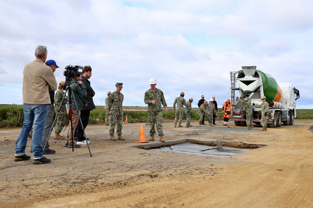Military personnel in U.S. Navy camouflage uniforms are conducting a concrete repair operation on what appears to be a damaged pavement surface, likely a runway or road. A concrete mixer truck is actively dispensing concrete into a prepared excavation while a media crew with a camera on a tripod documents the activity. One individual wearing a white hard hat appears to be in a supervisory or leadership role.