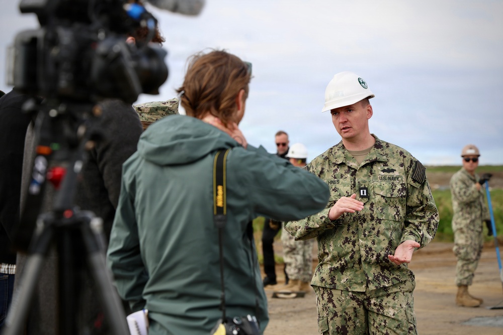 A U.S. Navy officer wearing a white hard hat and NWU Type III camouflage uniform is being interviewed by a civilian journalist with a camera crew present. The officer, identified by name tape as 'HINES' and wearing a Captain's rank insignia, appears to be speaking at an outdoor site. Additional military personnel and civilians in hard hats are visible in the background.