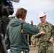 A U.S. Navy officer wearing a white hard hat and NWU Type III camouflage uniform is being interviewed by a civilian journalist with a camera crew present. The officer, identified by name tape as 'HINES' and wearing a Captain's rank insignia, appears to be speaking at an outdoor site. Additional military personnel and civilians in hard hats are visible in the background.
