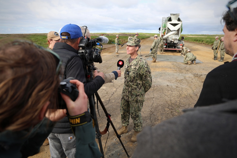 A U.S. Navy service member in camouflage uniform is being interviewed by a television news crew, identifiable by a microphone bearing a '4' logo, at an outdoor construction or road repair site. In the background, multiple military personnel are working alongside a concrete mixer truck on what appears to be a dirt or gravel road. The setting appears to be a flat, open terrain under overcast skies.
