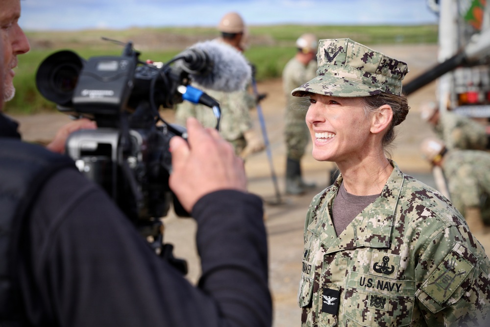 A U.S. Navy officer in NWU Type III camouflage uniform is being filmed by a civilian videographer during what appears to be an outdoor media interview. The officer, wearing a Captain's eagle insignia and various qualification badges, is smiling broadly at the camera. Additional Navy personnel are visible working in the background.