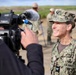 A U.S. Navy officer in NWU Type III camouflage uniform is being filmed by a civilian videographer during what appears to be an outdoor media interview. The officer, wearing a Captain's eagle insignia and various qualification badges, is smiling broadly at the camera. Additional Navy personnel are visible working in the background.