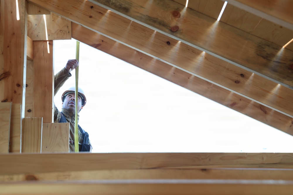 A construction worker wearing a white hard hat is taking measurements inside a wooden roof frame structure. The photo is taken from a low angle looking upward, revealing the triangular geometry of roof rafters and sheathing boards made of light-colored lumber. The worker appears to be using a measuring tape to assess dimensions within the framing.