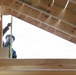 A construction worker wearing a white hard hat is taking measurements inside a wooden roof frame structure. The photo is taken from a low angle looking upward, revealing the triangular geometry of roof rafters and sheathing boards made of light-colored lumber. The worker appears to be using a measuring tape to assess dimensions within the framing.