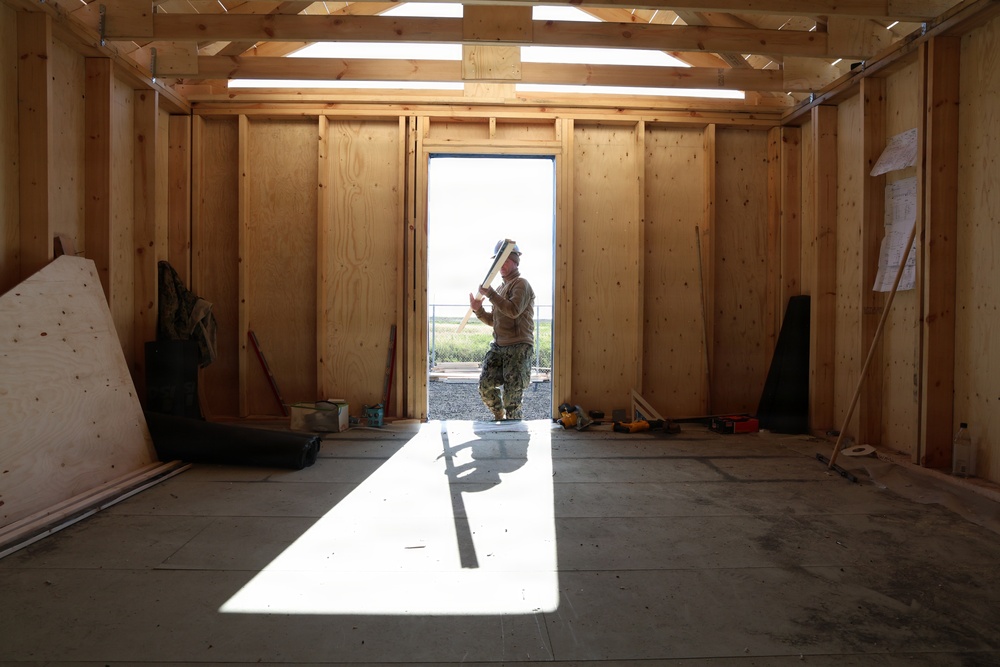 The image shows the interior of a building under construction with exposed wooden framing and a concrete floor. A single worker, appearing to wear military-style camouflage pants and a hard hat, stands in a doorway backlit by bright outdoor light. The space is partially finished with plywood sheathing on the walls and various construction tools and materials scattered around.