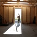 The image shows the interior of a building under construction with exposed wooden framing and a concrete floor. A single worker, appearing to wear military-style camouflage pants and a hard hat, stands in a doorway backlit by bright outdoor light. The space is partially finished with plywood sheathing on the walls and various construction tools and materials scattered around.