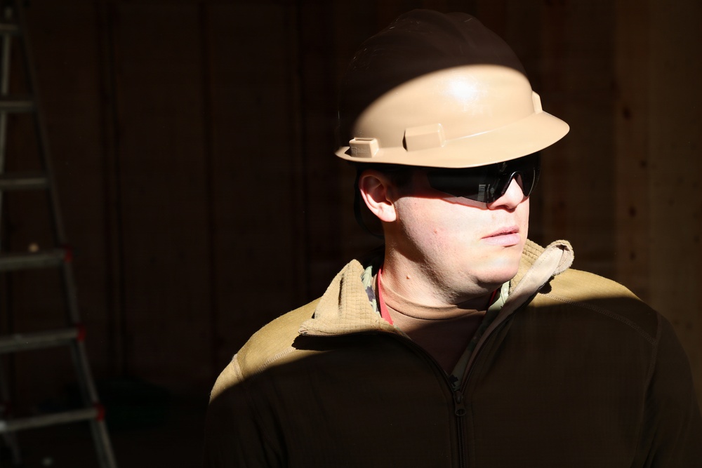 A person wearing a tan/beige hard hat and dark safety glasses is photographed in a construction or work environment. The subject is wearing a dark zip-up fleece jacket with a tan vest or body armor over it. The image features dramatic directional lighting that illuminates the subject's face and helmet against a dark wooden background.