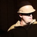 A person wearing a tan/beige hard hat and dark safety glasses is photographed in a construction or work environment. The subject is wearing a dark zip-up fleece jacket with a tan vest or body armor over it. The image features dramatic directional lighting that illuminates the subject's face and helmet against a dark wooden background.
