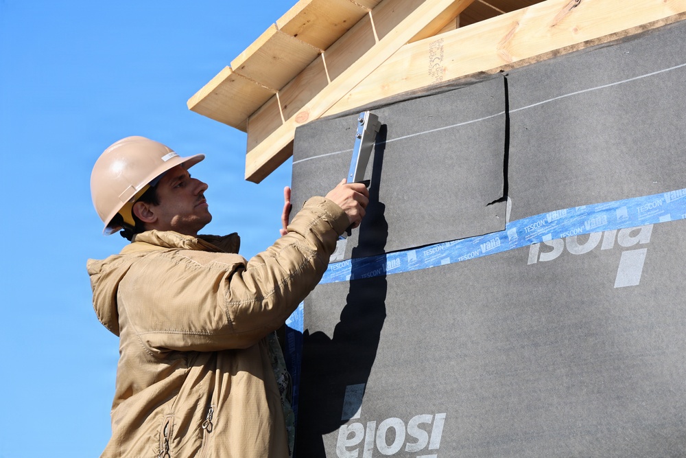 A construction worker wearing a hard hat and beige jacket is installing or securing a section of building wrap/weather barrier membrane on the exterior wall of a structure under construction. The wall is covered with a dark gray membrane branded 'Isola' with blue seam tape labeled 'TESCON Vana' applied at the joints. The wooden roof framing is visible at the top of the frame against a clear blue sky.