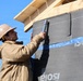 A construction worker wearing a hard hat and beige jacket is installing or securing a section of building wrap/weather barrier membrane on the exterior wall of a structure under construction. The wall is covered with a dark gray membrane branded 'Isola' with blue seam tape labeled 'TESCON Vana' applied at the joints. The wooden roof framing is visible at the top of the frame against a clear blue sky.