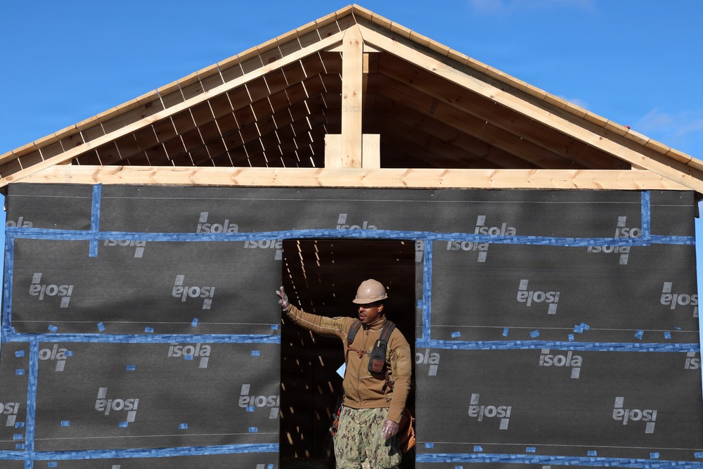 A construction worker wearing a hard hat and military-style camouflage pants stands in the doorway of a building under construction. The structure's exterior walls are covered with 'Isola' brand weather barrier/house wrap material secured with blue tape, while the roof framing (rafters and ridge board) is exposed wood. The scene appears to be an active residential or small building construction site under clear blue skies.