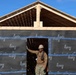A construction worker wearing a hard hat and military-style camouflage pants stands in the doorway of a building under construction. The structure's exterior walls are covered with 'Isola' brand weather barrier/house wrap material secured with blue tape, while the roof framing (rafters and ridge board) is exposed wood. The scene appears to be an active residential or small building construction site under clear blue skies.