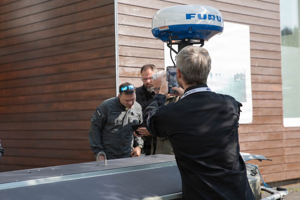 A group of people are gathered outdoors near a wooden building, examining what appears to be a boat or vessel equipped with a Furuno radar dome mounted on top. One person in the foreground is photographing the scene with a smartphone while wearing a lanyard/badge. Two other individuals are leaning over to inspect something on the vessel.