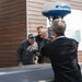 A group of people are gathered outdoors near a wooden building, examining what appears to be a boat or vessel equipped with a Furuno radar dome mounted on top. One person in the foreground is photographing the scene with a smartphone while wearing a lanyard/badge. Two other individuals are leaning over to inspect something on the vessel.