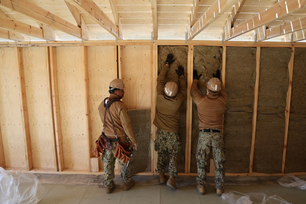 Three military personnel in camouflage uniforms and hard hats are installing insulation batts into a wood-framed wall structure inside a building under construction. Two workers are actively pressing insulation material into the wall cavities between studs while a third observes. The workers appear to be U.S. Navy Seabees or similar military construction unit based on their uniforms.