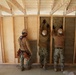 Three military personnel in camouflage uniforms and hard hats are installing insulation batts into a wood-framed wall structure inside a building under construction. Two workers are actively pressing insulation material into the wall cavities between studs while a third observes. The workers appear to be U.S. Navy Seabees or similar military construction unit based on their uniforms.