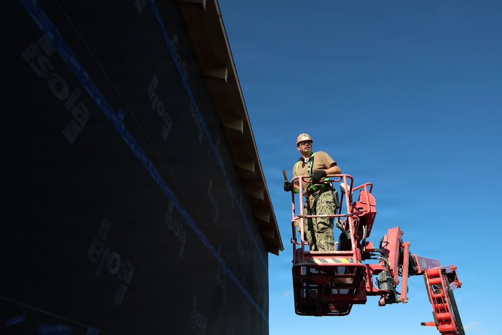 A construction worker is elevated on a red aerial lift (boom lift/cherry picker) alongside a large building wall covered in black weather barrier/house wrap material. The worker is wearing full safety gear including a hard hat, safety harness, and protective eyewear. The image is taken from a low angle against a clear blue sky, emphasizing the height and scale of the work being performed.
