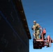 A construction worker is elevated on a red aerial lift (boom lift/cherry picker) alongside a large building wall covered in black weather barrier/house wrap material. The worker is wearing full safety gear including a hard hat, safety harness, and protective eyewear. The image is taken from a low angle against a clear blue sky, emphasizing the height and scale of the work being performed.
