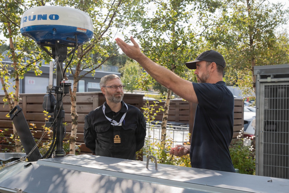 Two men are engaged in a discussion near a marine radar dome unit, likely branded 'Furuno', mounted on a mast. One man wearing a black uniform with rank insignia and a lanyard listens attentively while the other, in a black t-shirt and cap, gestures expressively with one hand. The setting appears to be an outdoor area near a marina or technical facility.