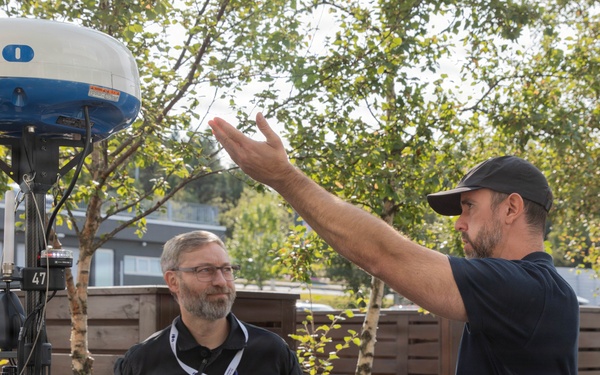 Two men are engaged in a discussion near a marine radar dome unit, likely branded 'Furuno', mounted on a mast. One man wearing a black uniform with rank insignia and a lanyard listens attentively while the other, in a black t-shirt and cap, gestures expressively with one hand. The setting appears to be an outdoor area near a marina or technical facility.